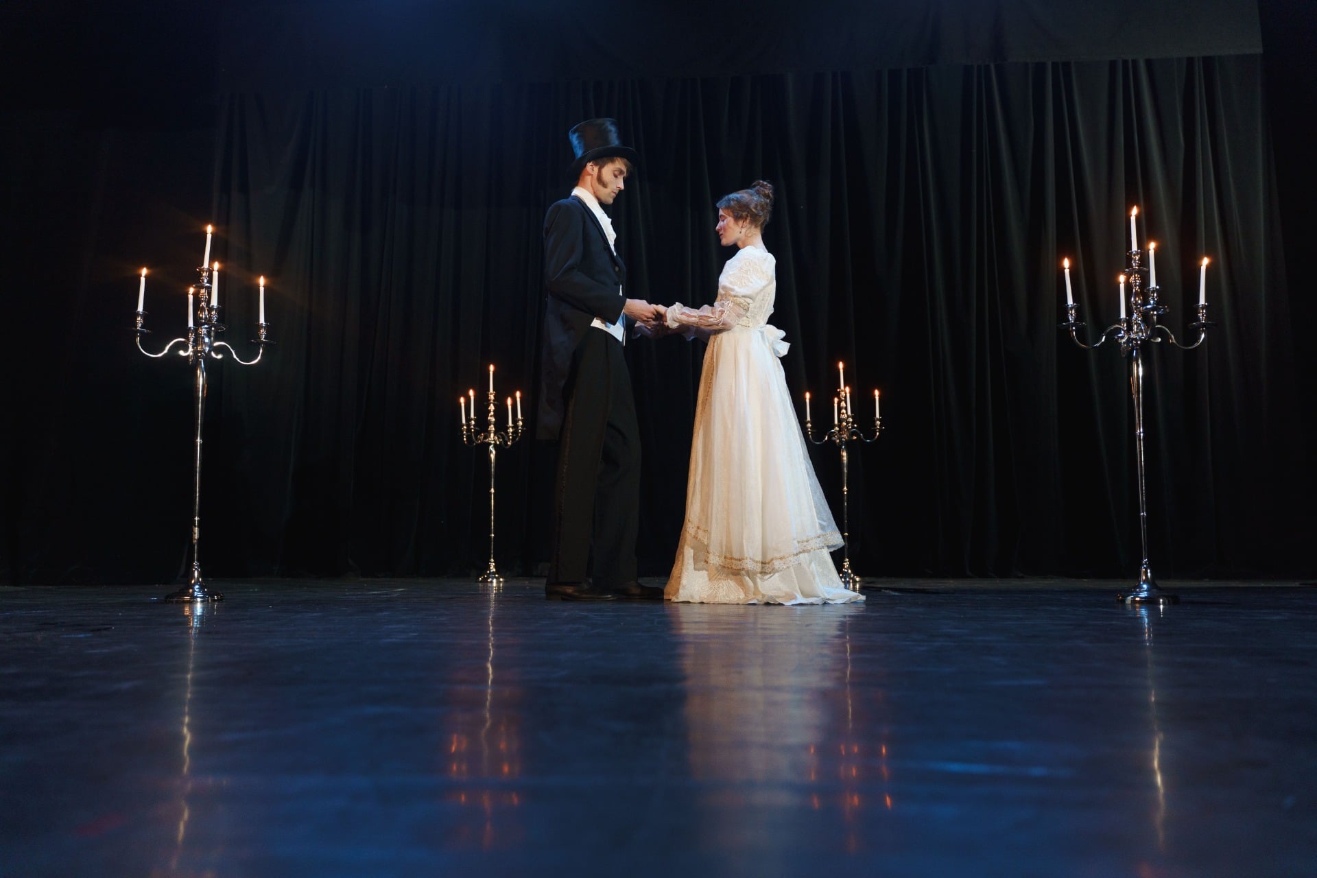 Casal em figurino de época de mãos dadas no palco, entre candelabros acesos